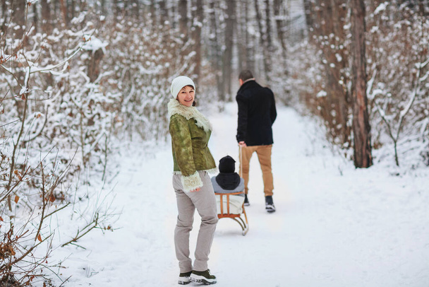 Cheerful parents walking behind glad boy with sleigh while spending time in snowy woods on winter day - Фото, изображение