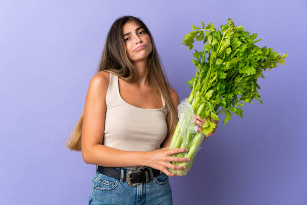 Mladá žena drží celer izolované na fialovém pozadí se smutným výrazem - Fotografie, Obrázek