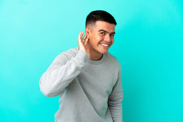 Young handsome man over isolated blue background listening to something by putting hand on the ear - Foto, imagen