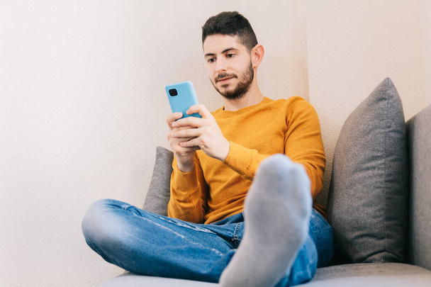 Nuori aikuinen mies hänen 30s valkoihoinen käyttäen matkapuhelinta sohvalla kotona, katsoen näytön onnellinen ja hauska - Valokuva, kuva