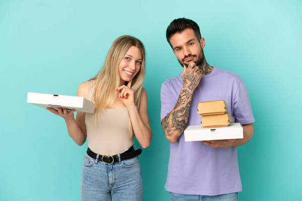 Пара держа пиццу и бургеры на изолированном синем фоне улыбаясь со сладким выражением лица - Фото, изображение