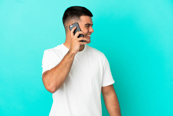 Jeune homme beau sur fond bleu isolé gardant une conversation avec le téléphone portable avec quelqu'un - Photo, image