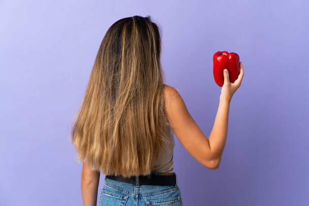 Young woman holding a pepper isolated on purple background in back position - Photo, Image