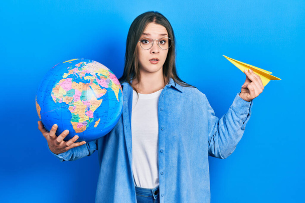 Young hispanic girl holding paper plane and world ball in shock face, looking skeptical and sarcastic, surprised with open mouth  - Photo, Image