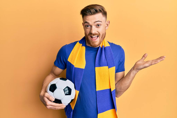 Junger rothaariger Mann Fußball-Hooligan mit Ball feiert Leistung mit glücklichem Lächeln und Siegerausdruck mit erhobener Hand  - Foto, Bild