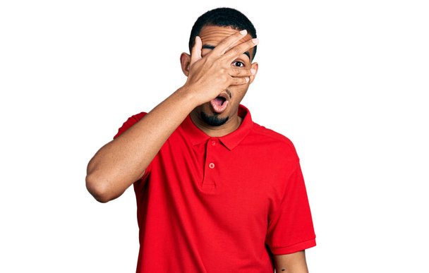 Fiatal afro-amerikai férfi alkalmi pólóban leselkedik sokk fedi arcát és szemét kézzel, néz keresztül ujjak félelem  - Fotó, kép