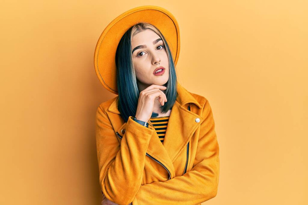 Νεαρό σύγχρονο κορίτσι φορώντας κίτρινο καπέλο και δερμάτινο μπουφάν με το χέρι στο πηγούνι σκεφτόμαστε την ερώτηση, σκεπτική έκφραση. Χαμογελώντας με ευγενικό πρόσωπο. έννοια της αμφιβολίας.  - Φωτογραφία, εικόνα