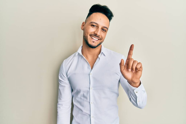 Fiatal arab férfi alkalmi ruhát visel mosolyog egy ötlettel vagy kérdéssel mutogatva ujjal felfelé boldog arccal, első számú  - Fotó, kép