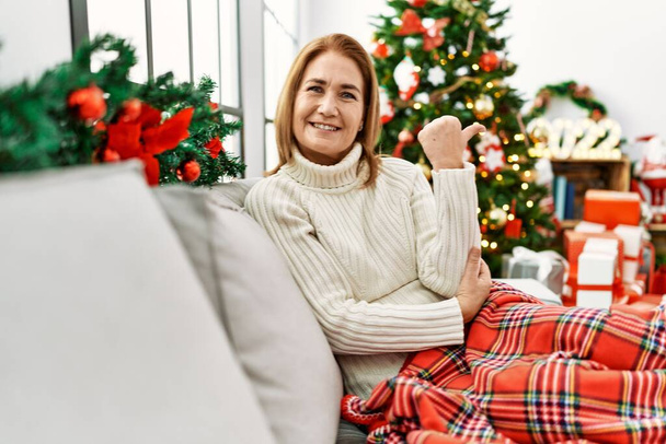 Женщина средних лет, сидящая на диване у рождественской елки, улыбаясь с счастливым лицом, глядя и указывая на сторону с большим пальцем вверх.  - Фото, изображение