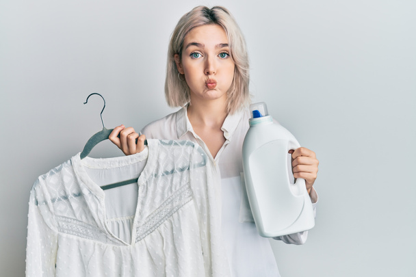 Jovencita rubia sosteniendo botella de detergente y camisa hinchando mejillas con cara divertida. boca inflada con aire, captación de aire.  - Foto, imagen