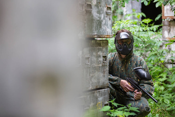 Πορτρέτο του νεαρού άνδρα με μάσκα προσώπου και όπλο στο παιχνίδι δράσης του paintball, προσομοιώνουν στρατιωτική μάχη χρησιμοποιώντας αεροβόλα όπλα για να πυροβολήσει κάψουλες του χρώματος σε κάθε άλλο - Φωτογραφία, εικόνα
