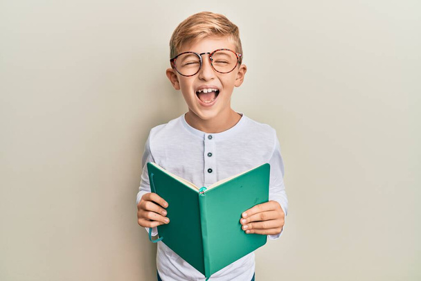 Маленький білий хлопчик читає книгу в окулярах, посміхається і сміється важко вголос, тому що смішний божевільний жарт
.  - Фото, зображення