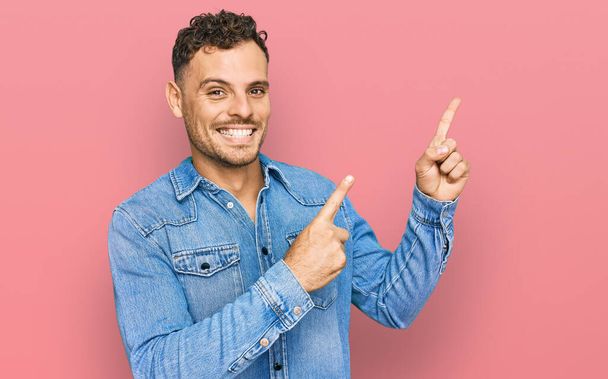 Junger hispanischer Mann in lässiger Jeansjacke lächelt und blickt mit zwei Händen und Fingern zur Seite in die Kamera.  - Foto, Bild