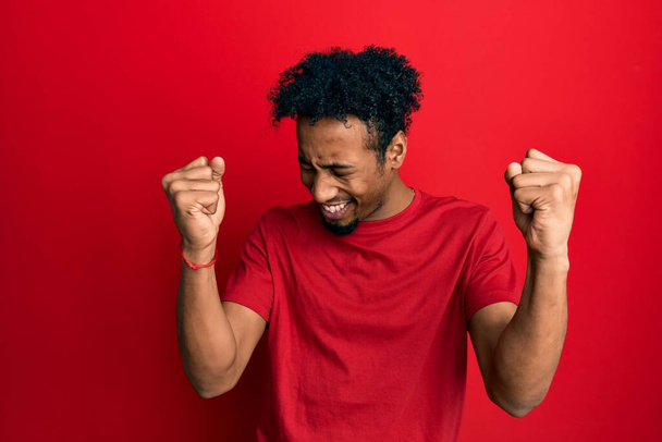 Νεαρός Αφροαμερικάνος με γενειάδα που φοράει κόκκινο μπλουζάκι πολύ χαρούμενος και ενθουσιασμένος που κάνει τη χειρονομία του νικητή με τα χέρια σηκωμένα, χαμογελώντας και ουρλιάζοντας για επιτυχία. έννοια εορτασμού.  - Φωτογραφία, εικόνα