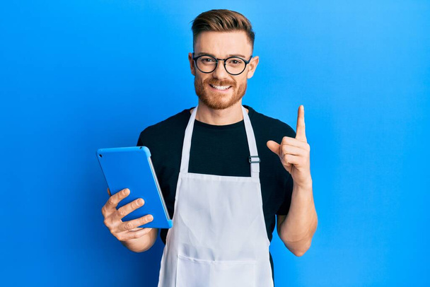 Young redhead man wearing professional apron using touchpad smiling with an idea or question pointing finger with happy face, number one  - Photo, Image