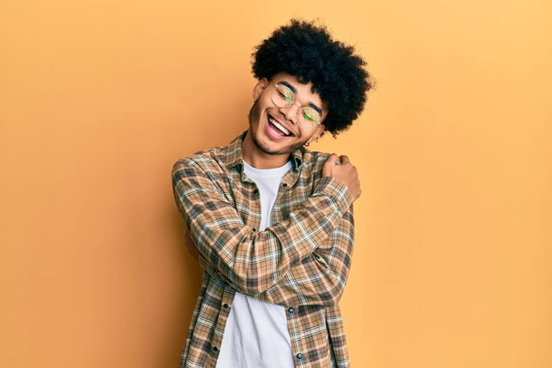 Young african american man with afro hair wearing casual clothes hugging oneself happy and positive, smiling confident. self love and self care  - Фото, изображение