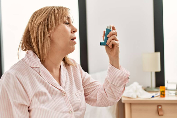 Orta yaşlı bir kadın astım ilacı kullanıyor. Evdeki yatakta oturuyor.. - Fotoğraf, Görsel