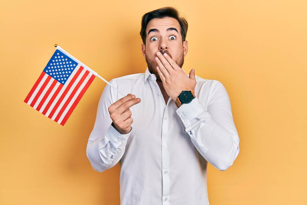 Bel homme caucasien avec barbe tenant drapeau des États-Unis couvrant la bouche avec la main, choqué et effrayé par l'erreur. expression surprise  - Photo, image