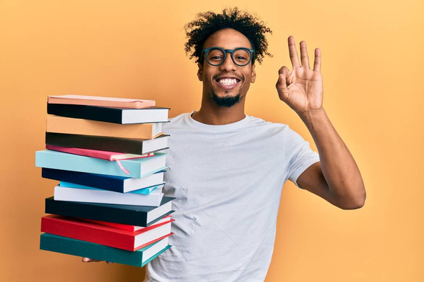 Mladý africký Američan s plnovousem drží hromadu knih dělá v pořádku znamení s prsty, usmívá přátelsky gestikulující vynikající symbol  - Fotografie, Obrázek