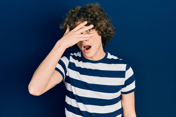 Handsome young man wearing casual striped t shirt peeking in shock covering face and eyes with hand, looking through fingers with embarrassed expression.  - 写真・画像