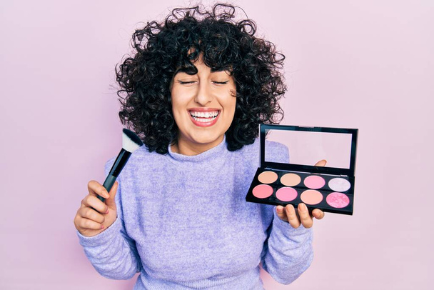 Young middle east woman holding makeup brush and blush smiling and laughing hard out loud because funny crazy joke.  - Photo, Image