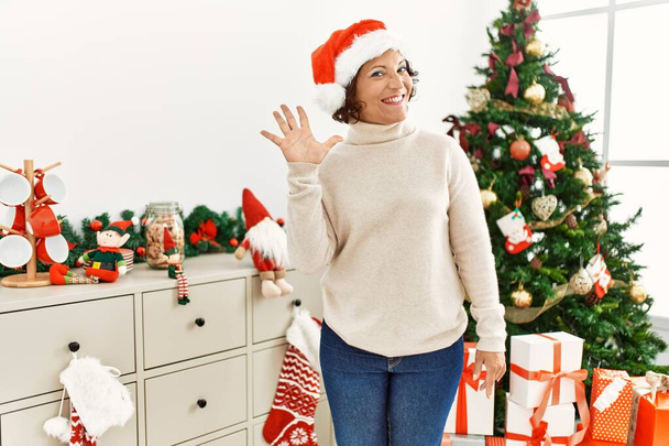 Mujer hispana de mediana edad de pie junto al árbol de Navidad mostrando y señalando hacia arriba con los dedos número cinco mientras sonríe confiada y feliz.  - Foto, imagen