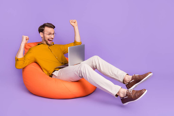 Portrait of attractive cheerful guy sitting on chair using laptop rejoicing isolated over violet purple color background - Фото, зображення