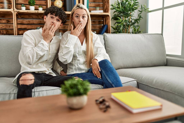 Jovem casal bonito sentado no sofá em casa cobrindo a boca com a mão, chocado e com medo de erro. expressão surpreendida  - Foto, Imagem