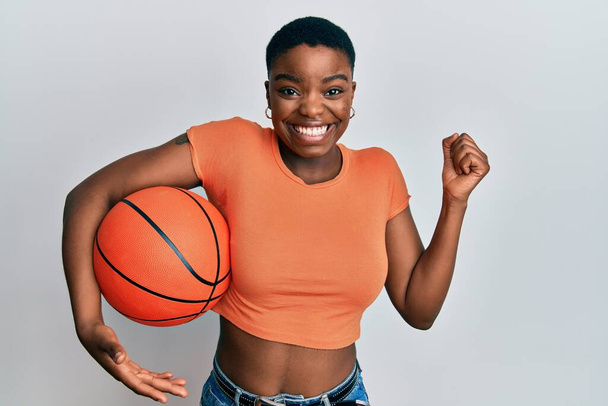 Jeune femme afro-américaine tenant ballon de basket criant fier, célébrant la victoire et le succès très excité avec le bras levé  - Photo, image