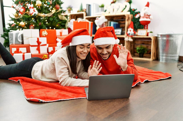 Jovem casal hispânico tendo videochamada usando laptop deitado no chão pela árvore de natal em casa. - Foto, Imagem