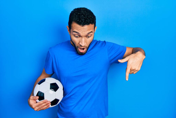 Ισπανόφωνος άνδρας με γένια κρατώντας μπάλα ποδοσφαίρου δείχνει προς τα κάτω με τα δάχτυλα δείχνει διαφήμιση, έκπληκτος πρόσωπο και ανοιχτό στόμα  - Φωτογραφία, εικόνα
