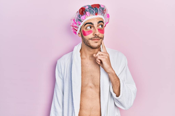 Giovane uomo ispanico che indossa accappatoio e cuffia da doccia utilizzando borse occhi patch viso serio pensando alla domanda con mano sul mento, riflessivo sull'idea confusa  - Foto, immagini