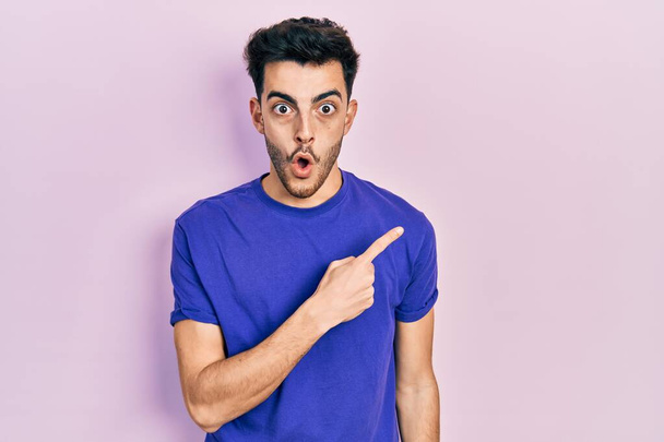 Joven hombre hispano vistiendo camiseta casual sorprendido señalando con el dedo hacia un lado, boca abierta expresión asombrada.  - Foto, Imagen