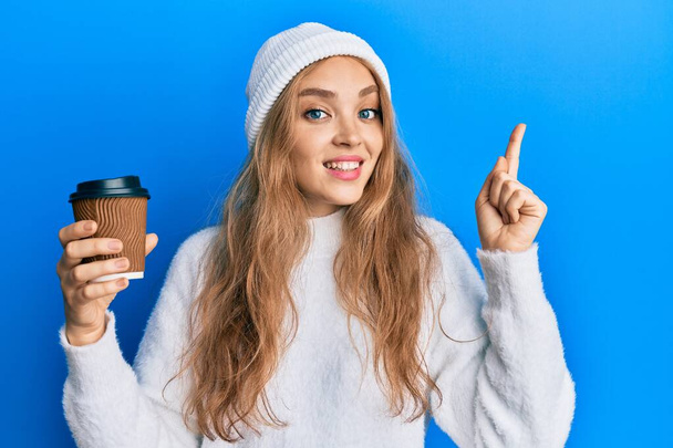 Mooie blonde blanke vrouw drinken van een take away kopje koffie glimlachen met een idee of vraag wijzend vinger met gelukkig gezicht, nummer een  - Foto, afbeelding