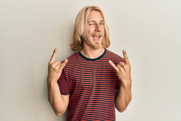 Caucasian young man with long hair doing rock gesture winking looking at the camera with sexy expression, cheerful and happy face.  - Foto, imagen