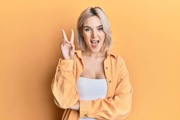 Young blonde girl wearing casual clothes smiling with happy face winking at the camera doing victory sign. number two.  - 写真・画像