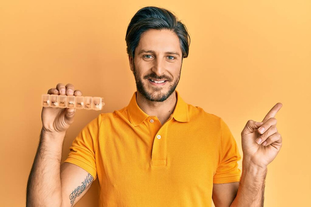 Молодой латиноамериканец, держащий в руках органайзер таблеток, счастливо улыбающийся, указывая рукой и пальцем в сторону  - Фото, изображение