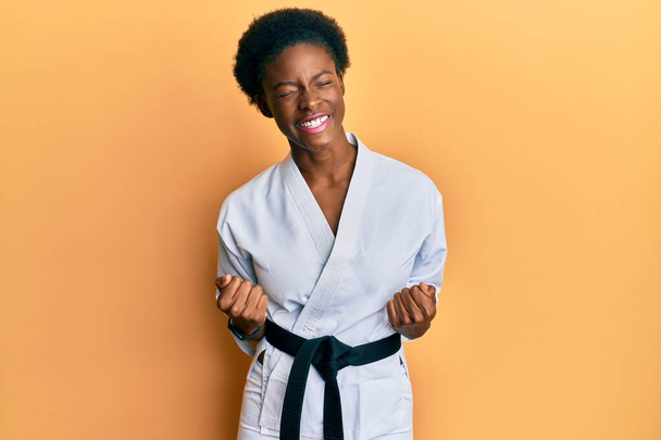 Nuori afrikkalainen amerikkalainen tyttö yllään karate kimono ja musta vyö erittäin onnellinen ja innoissaan tekee voittaja ele kädet ylhäällä, hymyillen ja huutaen menestystä. juhla käsite.  - Valokuva, kuva