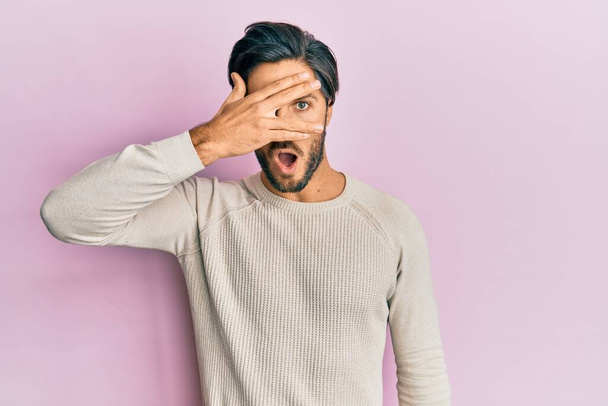 Jonge Spaanse man draagt casual winter trui gluren in shock bedekking gezicht en ogen met de hand, kijkend door vingers met beschaamde uitdrukking.  - Foto, afbeelding