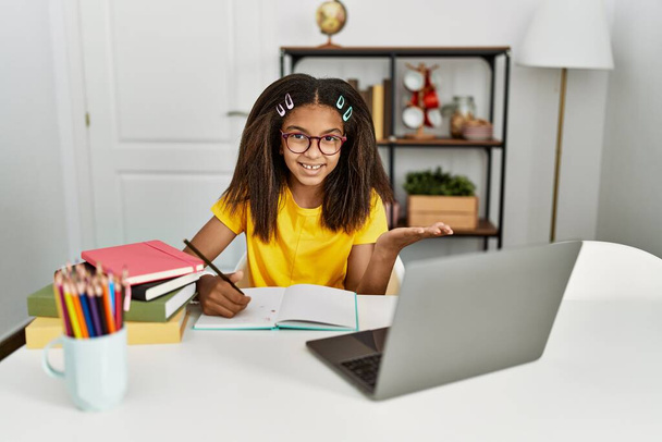 jong Afrikaans amerikaans meisje doen huiswerk thuis glimlachend vrolijke presentatie en wijzen met palm van de hand kijken naar de camera.  - Foto, afbeelding