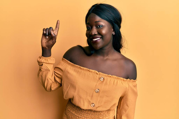 Jeune femme africaine portant des vêtements décontractés souriant avec une idée ou une question pointant du doigt avec un visage heureux, numéro un  - Photo, image