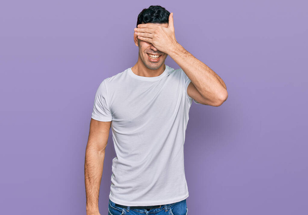 Handsome hispanic man wearing casual white t shirt smiling and laughing with hand on face covering eyes for surprise. blind concept.  - Foto, imagen