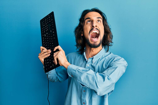 Jeune homme hispanique tenant le clavier en colère et fou hurlant frustré et furieux, criant de colère levant les yeux.  - Photo, image