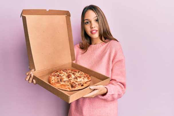 Mladá brunetka, držící italskou pizzu, se uvolnila s vážným výrazem ve tváři. jednoduchý a přirozený pohled do kamery.  - Fotografie, Obrázek