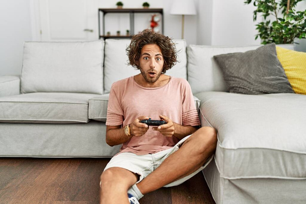 Νεαρός ισπανόφωνος άνδρας παίζει video game κρατώντας χειριστήριο στο σπίτι φοβισμένος και έκπληκτος με ανοιχτό το στόμα για έκπληξη, δυσπιστία πρόσωπο  - Φωτογραφία, εικόνα