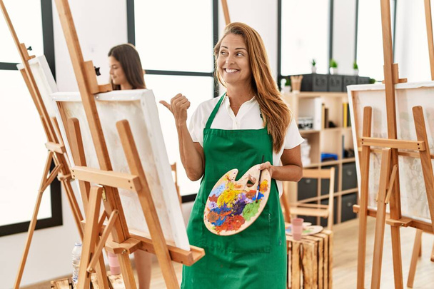 Giovane donna di mezza età ispanica in classe d'arte che punta il pollice verso l'alto di lato sorridente felice con la bocca aperta  - Foto, immagini