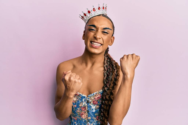 Homem hispânico vestindo maquiagem e cabelos longos vestindo coroa princesa gritando orgulhoso, celebrando vitória e sucesso muito animado com braço levantado  - Foto, Imagem