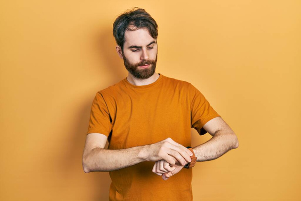 Homem caucasiano com barba vestindo casual camiseta amarela verificando o tempo no relógio de pulso, relaxado e confiante  - Foto, Imagem