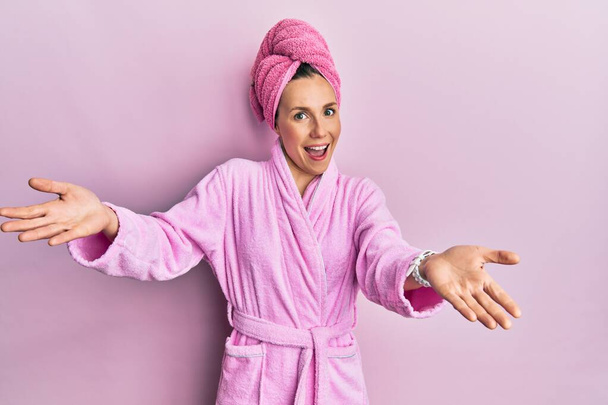 Jonge blonde vrouw met douche handdoekkap en badjas glimlachend vrolijk aanbieden van handen geven van hulp en acceptatie.  - Foto, afbeelding