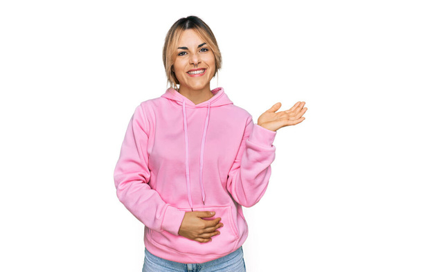 Young caucasian woman wearing casual sweatshirt smiling cheerful presenting and pointing with palm of hand looking at the camera.  - Photo, Image